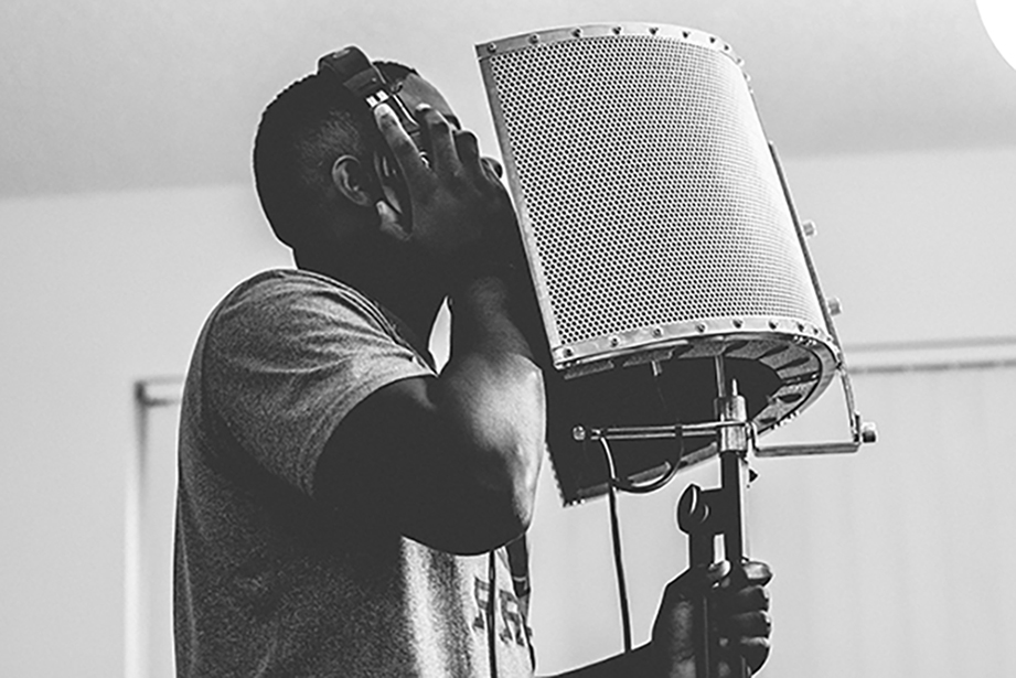 a man standing, singing into a recording micrphone in a studio room. 